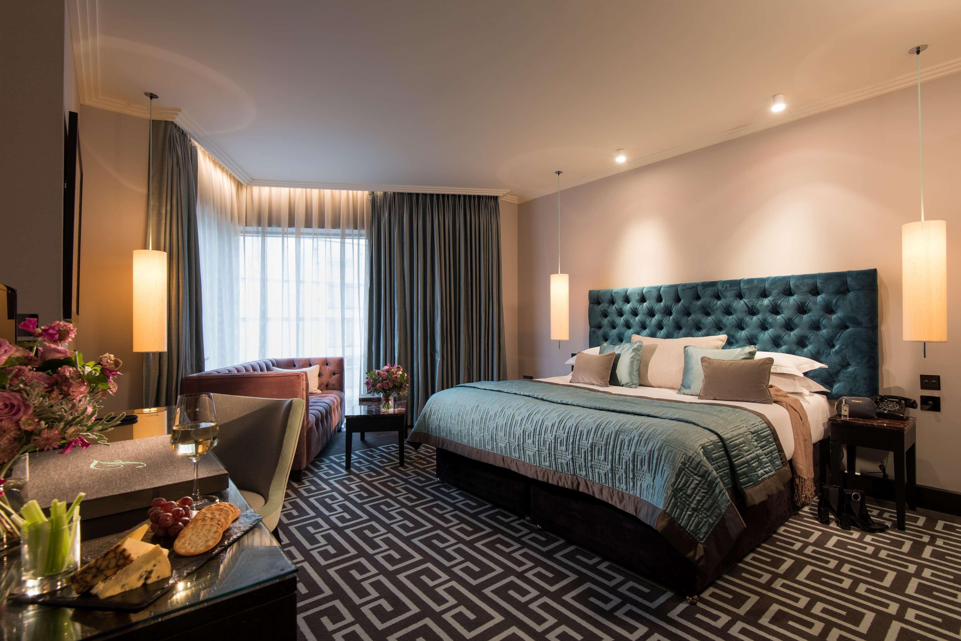 The Fitzwilliam Hotel Belfast Exteriér fotografie The photo depicts a stylish hotel room. It features a large bed with a tufted teal headboard, flanked by two wall sconces providing soft lighting. The bedding is neatly arranged with accent pillows. To one side, there is a small seating area with a c